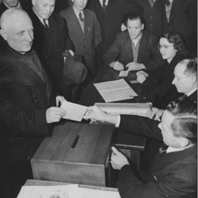 Das Bild zeigt die Stimmabgabe des Bürgermeisters Wilhelm Arnoul bei der Gemeinderatswahl am 27. Januar 1946 im Wahllokal in der Westendhalle. Zu sehen sind daneben v.l.n.r.: Georg Koser, Adolf Bauer, Philipp Wiemer, Adam Ebner, August Geiß (sitzend), Dr. Juliane Girardi-Vogt, Heinrich Zimbrich, Unbekannt, Fotograf: Paul Zehner, Offenbach]. Quelle: Stadtarchiv NI F 2.3 Nr. 49 Bld. 1